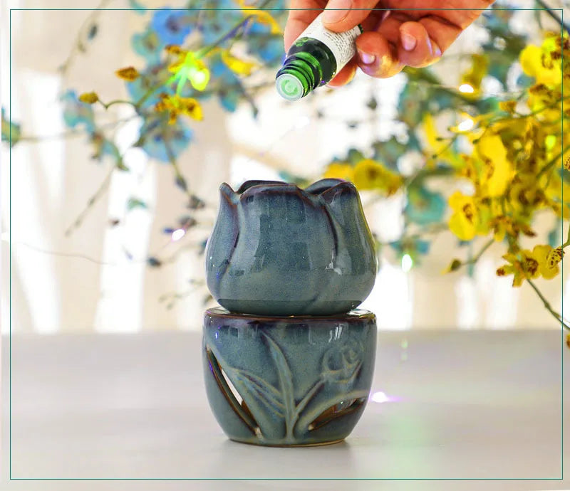 Ceramic lotus-shaped diffuser with a hand holding a green essential oil bottle against a blurred floral background. tulip ceramic essential oil incense burner, candle-powered aroma diffuser for home fragrance and calming boho home décor