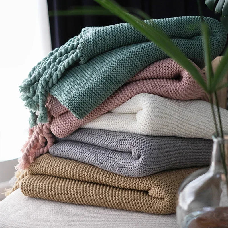 Stack of colorful knitted blankets on a surface with a plant in the foreground.