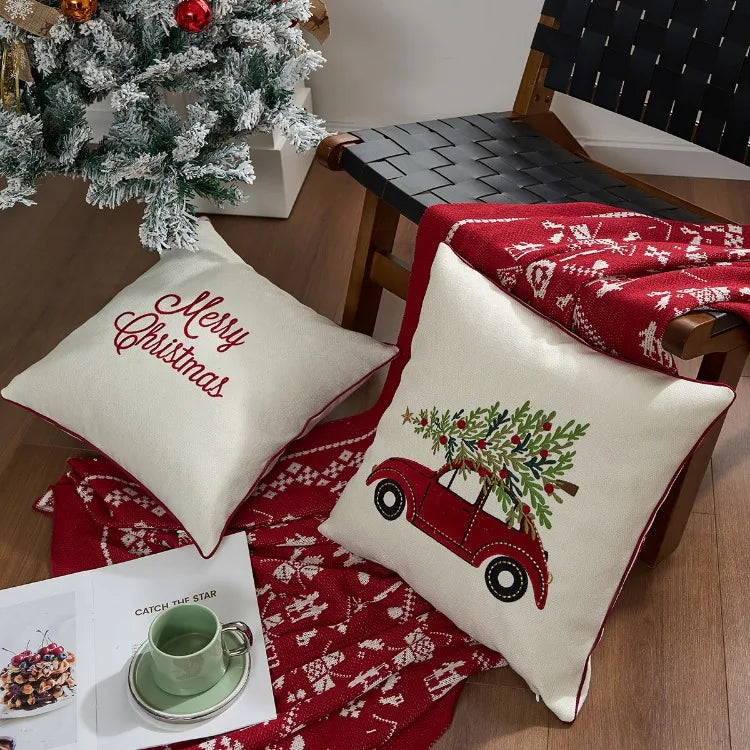 Decorative pillows with Christmas designs on a table with a red blanket and small tree.