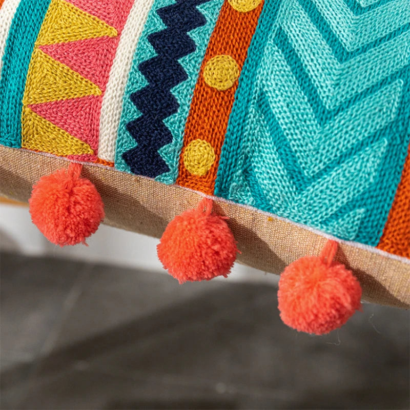 Colorful knitted fabric with geometric patterns and orange pom-poms on a neutral background. Bohemian embroidered throw pillow cover with geometric design, cotton-blend cushion for sofa, bed, or modern home decor.
