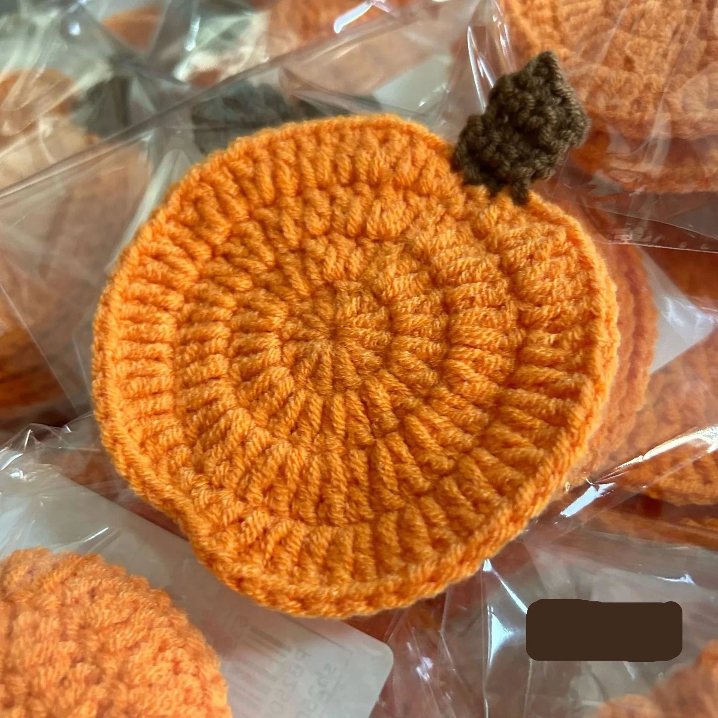 Orange crocheted item on a clear plastic background. Handmade crochet pumpkin coaster in cotton, Halloween table decor for mugs and drinks, festive DIY autumn party accessory