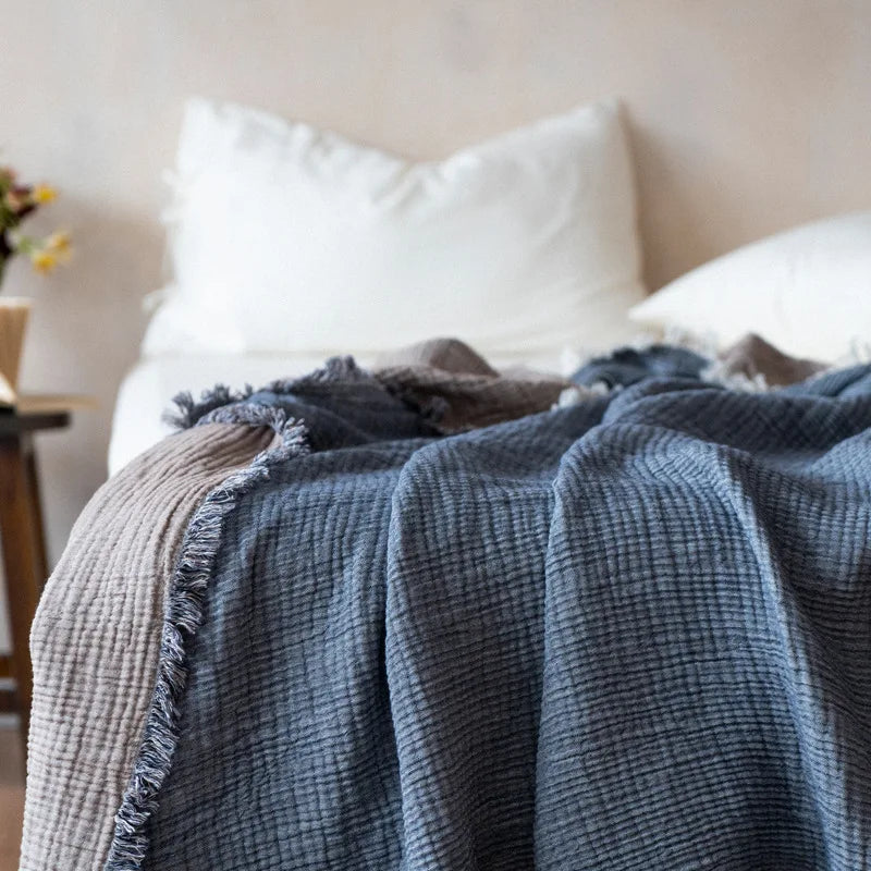 Blue textured blanket draped over a bed with white pillows. Maillard cotton gauze blanket with tassels, four-layer muslin throw for sofa, bed, and boho home decor