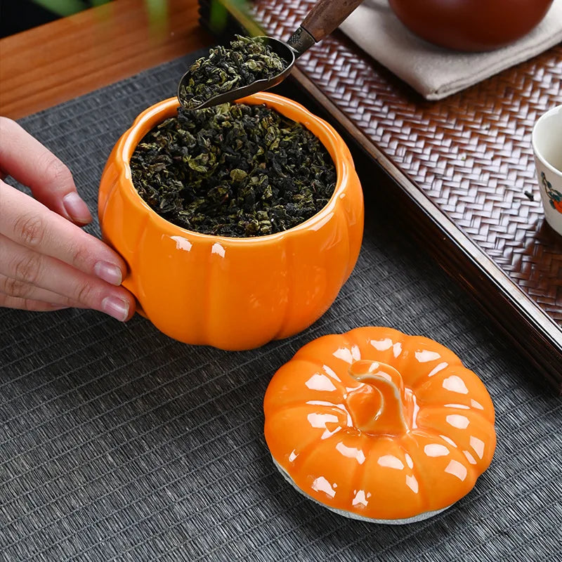 Orange pumpkin-shaped tea canister with lid on a textured surface, filled with green tea leaves. Ceramic pumpkin candy jar with lid, decorative fall tea jar or wedding gift container for home or halloween holiday decor.