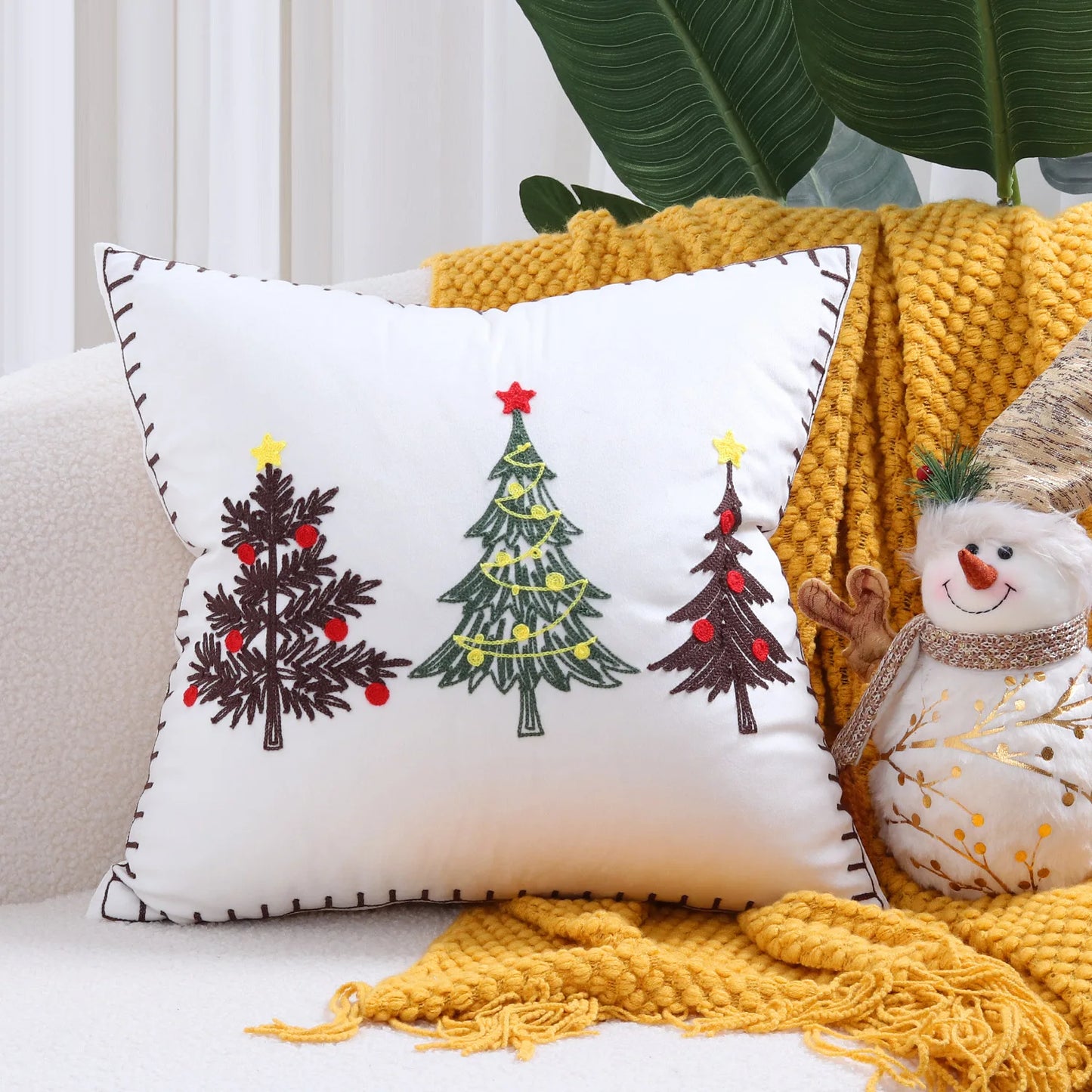 Decorative pillow with Christmas tree design on a couch with a yellow blanket and snowman toy.