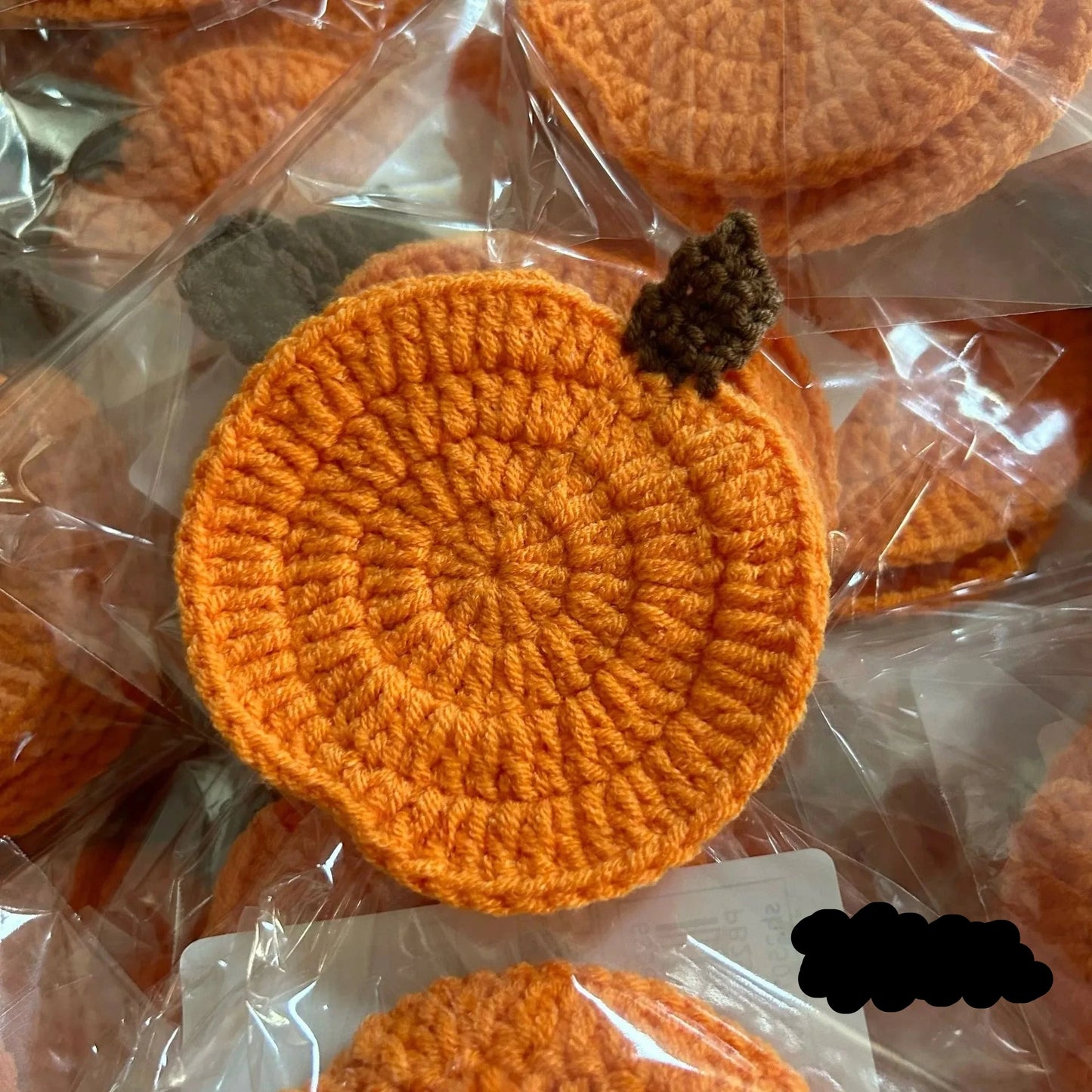 Orange crocheted items on clear plastic packaging. Handmade crochet pumpkin coaster in cotton, Halloween table decor for mugs and drinks, festive DIY autumn party accessory