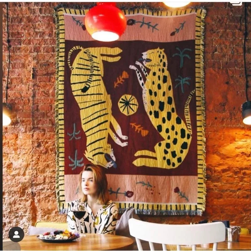 Woman sitting at a table with a colorful tapestry featuring leopards on the wall behind her. Leopard design boho home decor blanket, decorative woven tapestry for living room or bedroom