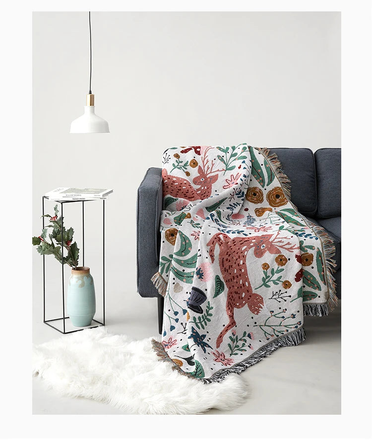 Floral and animal patterned blanket draped over a gray sofa with a white wall and decorative items in the background. Deer forest jacquard woven blanket with floral and woodland tapestry design – boho throw for sofa or bed, soft cotton blend decorative bedspread for cozy home décor.
