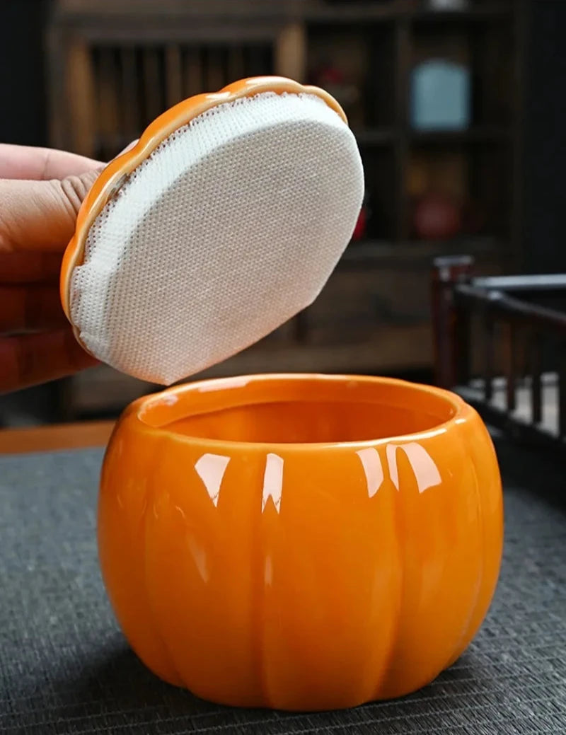 Orange ceramic tea canister with a lid being held open, on a dark surface. Ceramic pumpkin candy jar with lid, decorative fall tea jar or wedding gift container for home or halloween holiday decor.