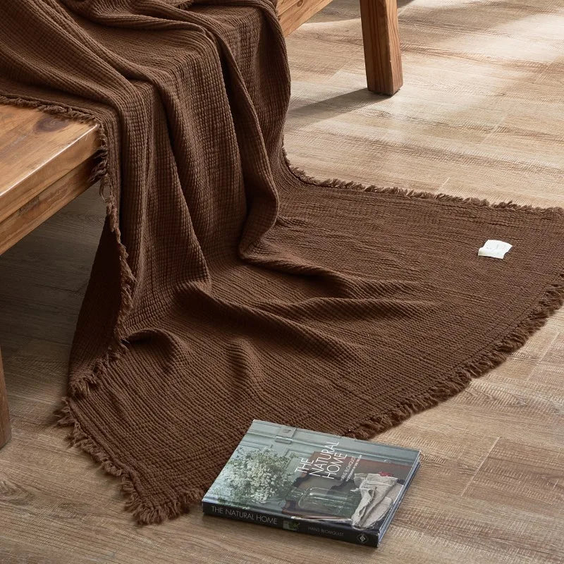 Brown textured blanket draped over a wooden bench on a wooden floor with a book underneath. Maillard cotton gauze blanket with tassels, four-layer muslin throw for sofa, bed, and boho home decor