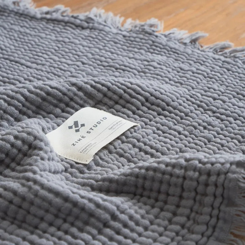 Gray textured blanket with a label on a wooden surface Maillard cotton gauze blanket with tassels, four-layer muslin throw for sofa, bed, and boho home decor