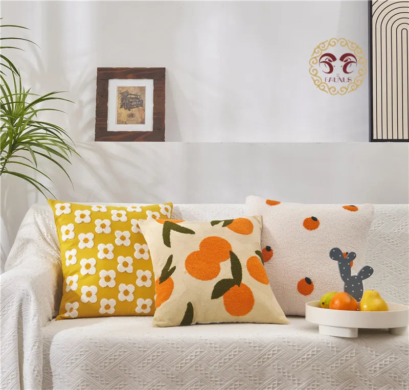 Decorative pillows with floral and fruit patterns on a sofa, with a plant and framed picture in the background.