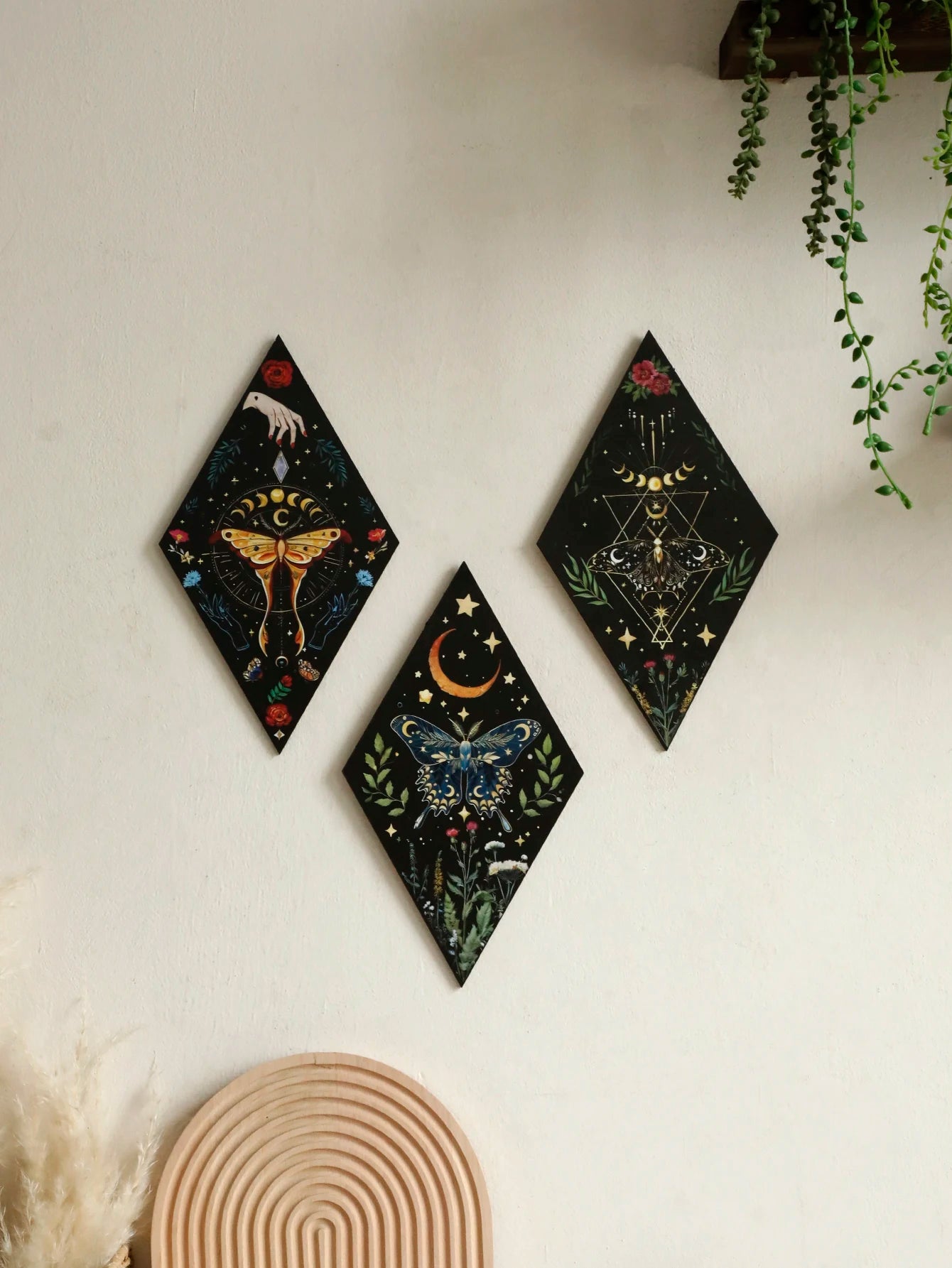 Three diamond-shaped wall art pieces with celestial designs on a white wall. Rustic colorful wood wall art panel with moon, stars, and butterfly engraving, displayed above a sofa in a boho-styled living room