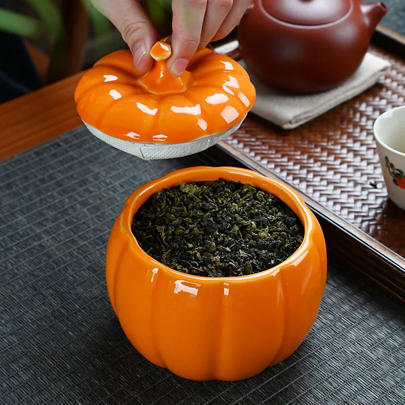 Orange ceramic tea canister with lid being opened, containing green tea leaves, on a dark surface. Ceramic pumpkin candy jar with lid, decorative fall tea jar or wedding gift container for home or halloween holiday decor.