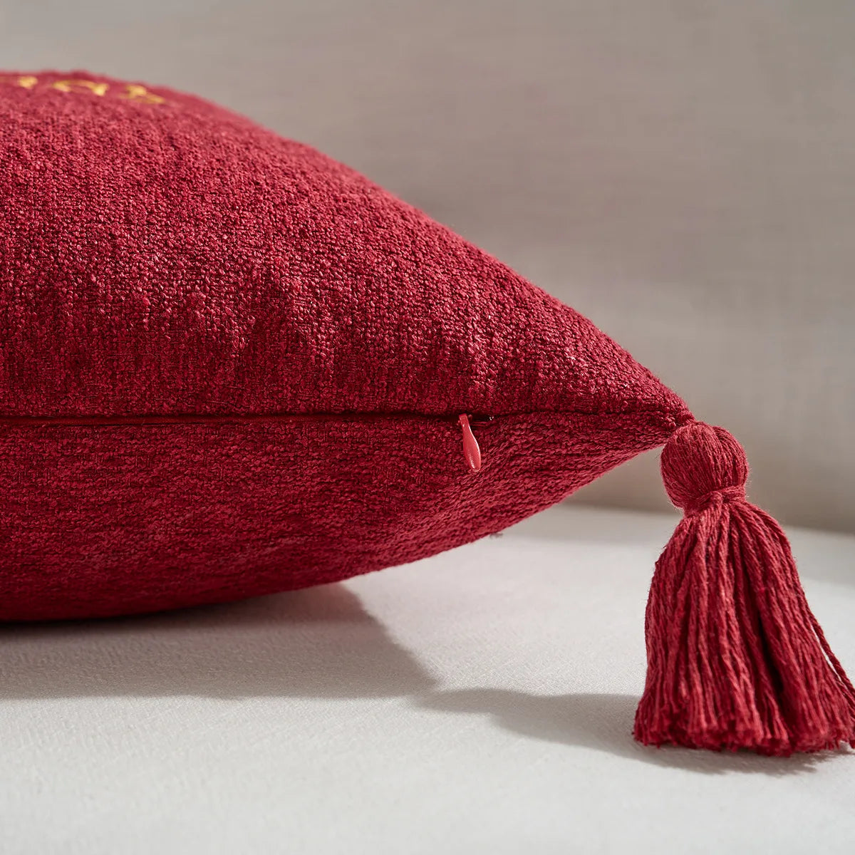 Red textured pillow with a tassel on a neutral background. Christmas gold letter embroidered pillow cover with “Holly Jolly” and snowflake design, soft red and white chenille cushion case for sofa or living room holiday décor.