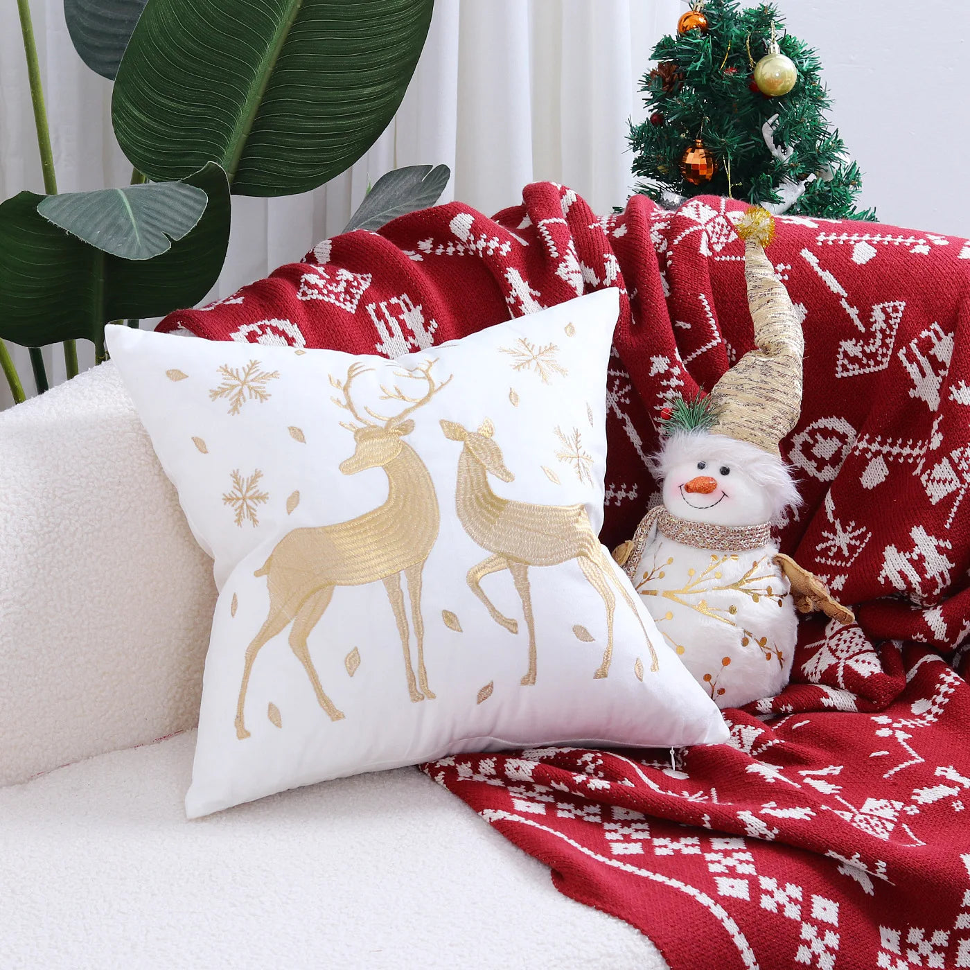 Decorative pillow with reindeer design on a couch with a red blanket and Christmas tree in the background.