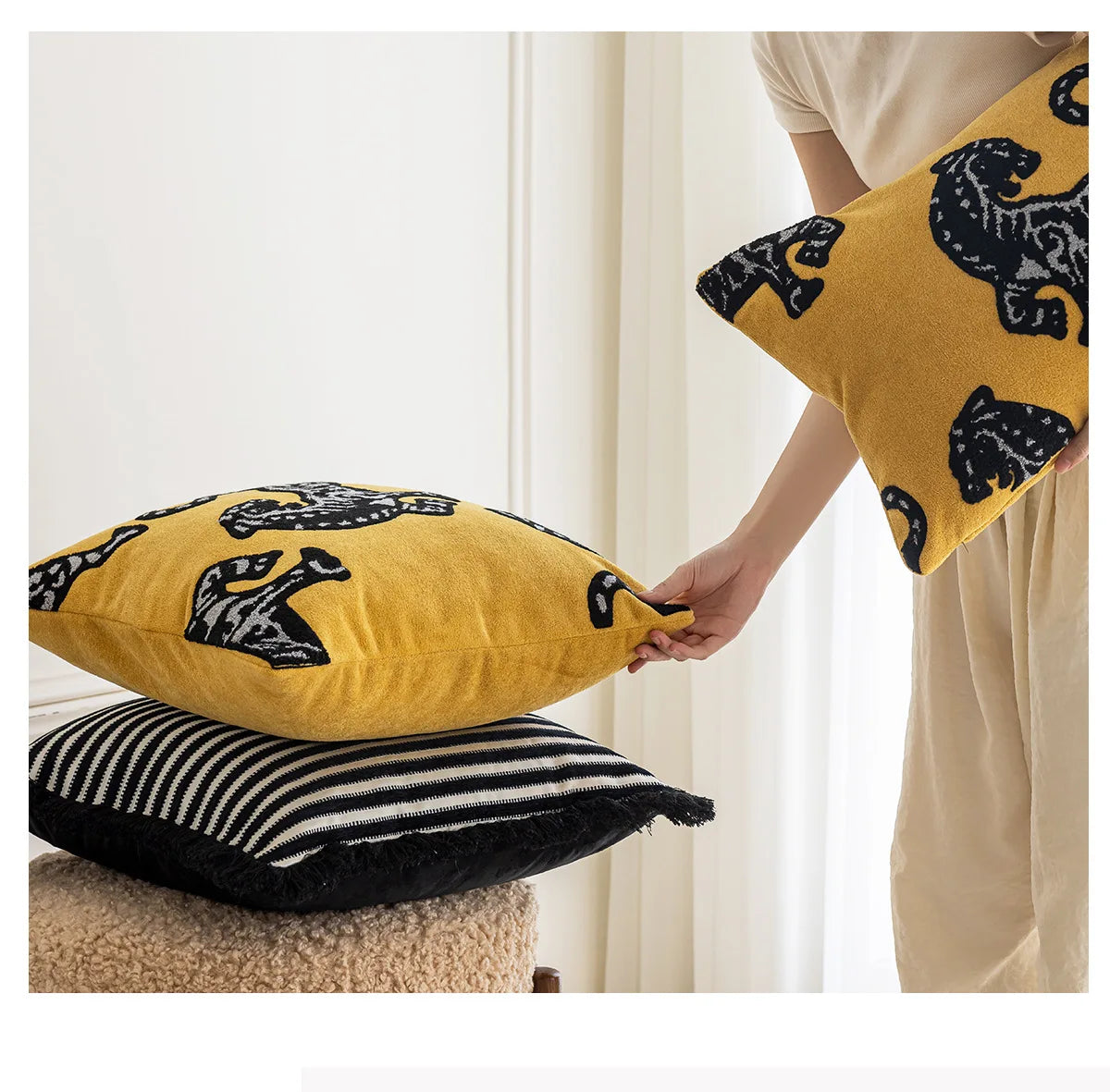 Person holding a yellow pillow with black patterns next to two other pillows on a surface. Modern minimalist embroidered pillow cover for living room decor.