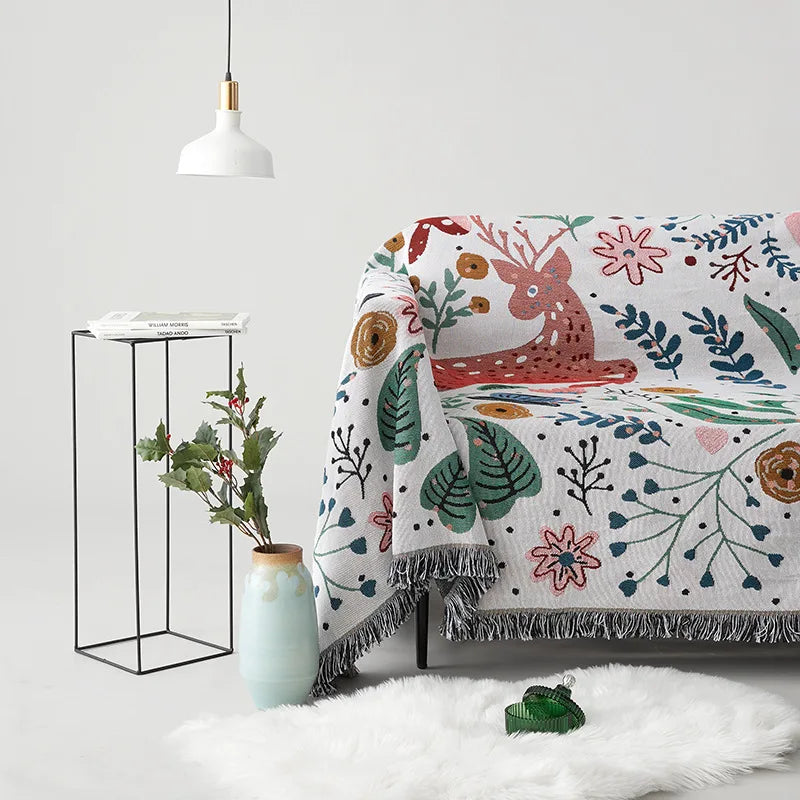 Decorative blanket with floral and deer pattern draped over a chair, with a vase and plant in the background. Deer forest jacquard woven blanket with floral and woodland tapestry design – boho throw for sofa or bed, soft cotton blend decorative bedspread for cozy home décor.