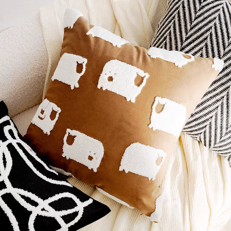 Decorative pillow with white sheep patterns on a brown background, surrounded by other pillows.