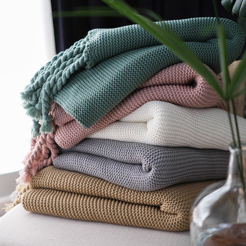 Stack of colorful boho knitted blankets on a surface with a plant in the foreground.