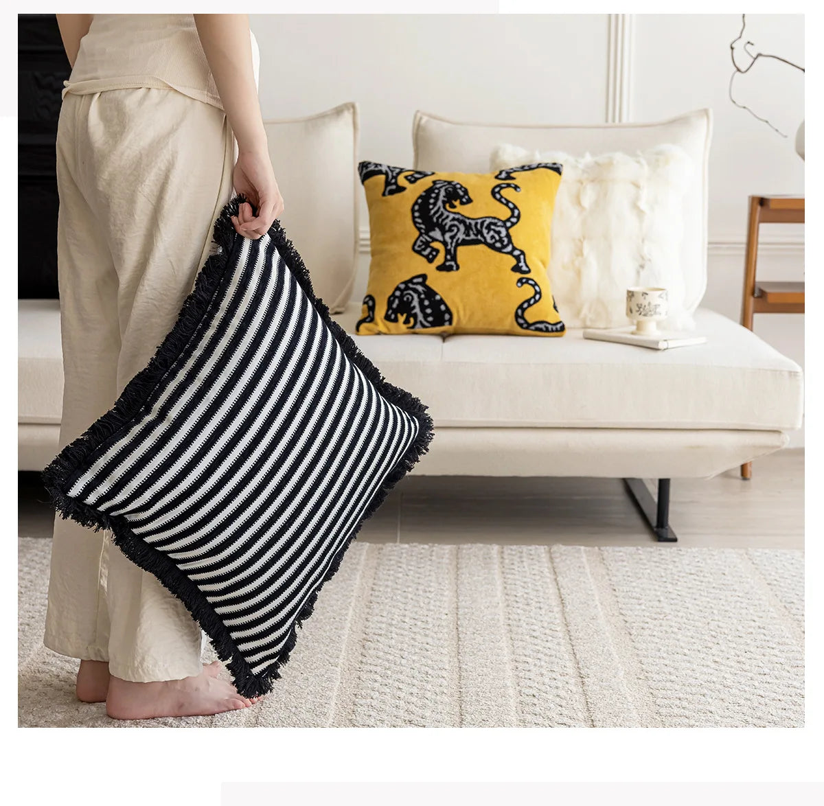 Person holding a black and white striped pillow with a couch and other pillows in the background. Modern minimalist embroidered pillow cover for living room decor.