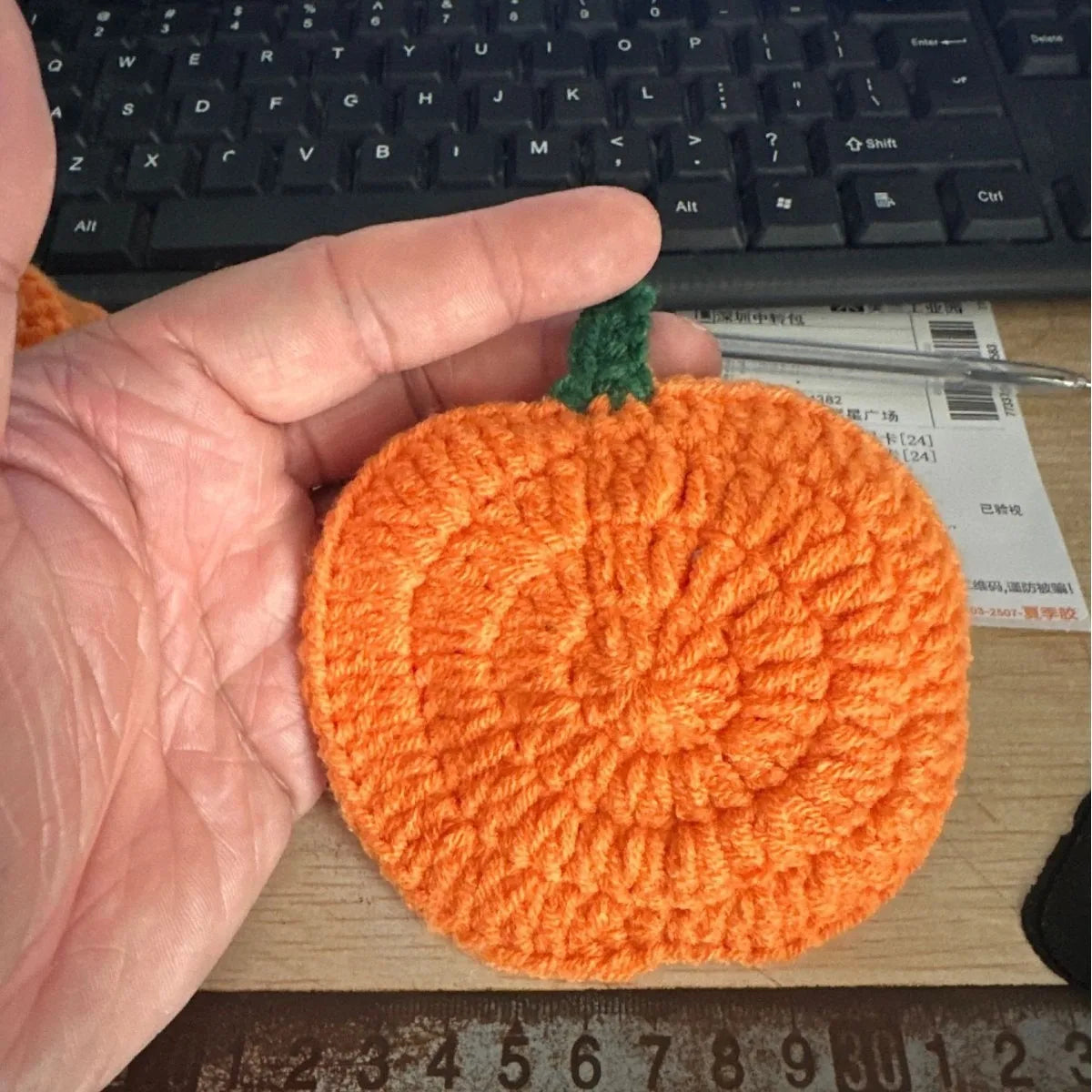 Hand holding a small orange crocheted pumpkin with a green stem, on a desk with a keyboard and paper in the background. Handmade crochet pumpkin coaster in cotton, Halloween table decor for mugs and drinks, festive DIY autumn party accessory