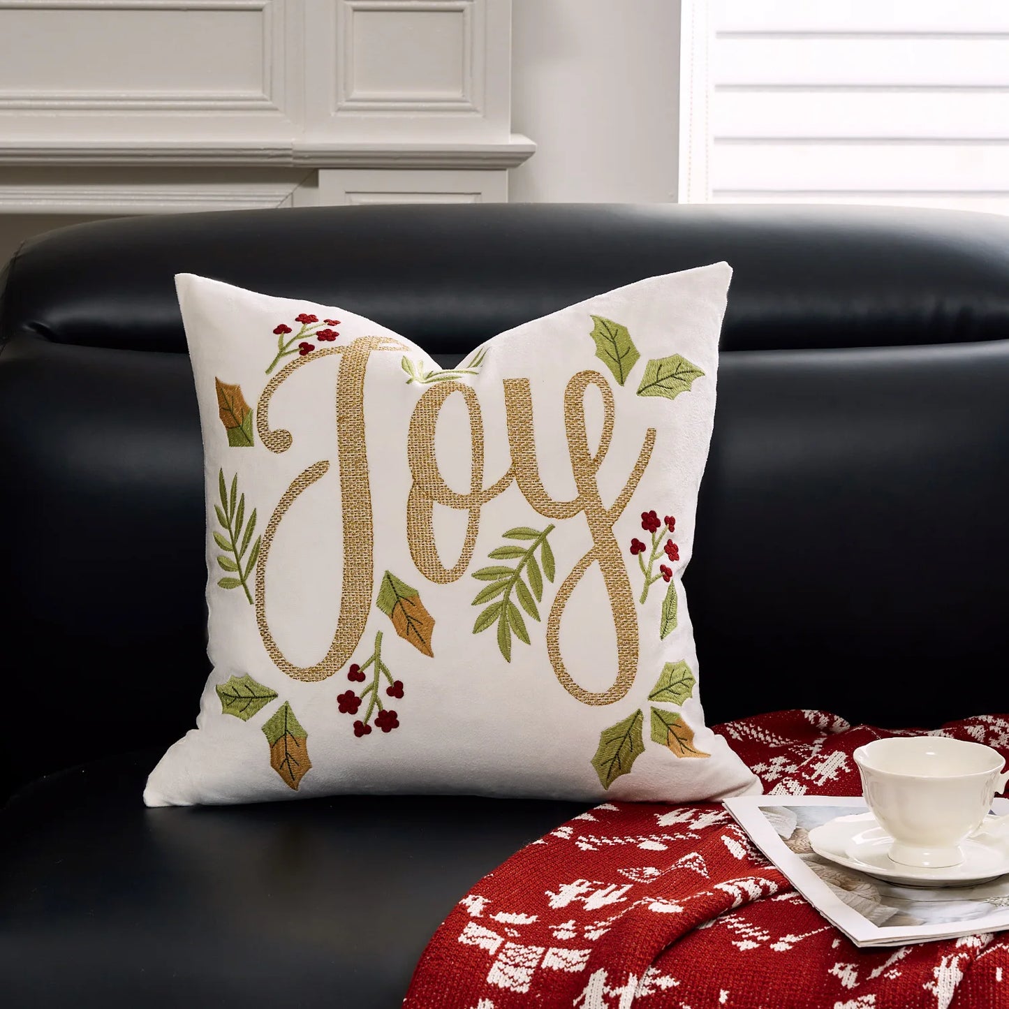 Decorative pillow with 'Joy' text on a black couch with a red blanket and white cup.