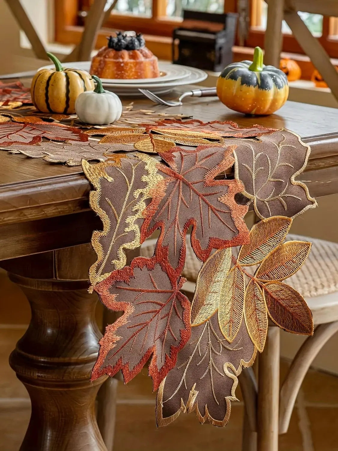 Decorative leaf-patterned table runner on a wooden table with pumpkins and a cake.