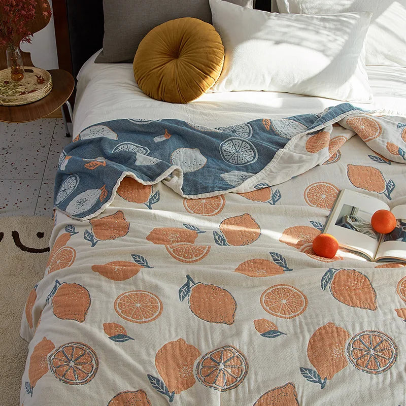 Bed with a decorative blanket featuring orange patterns and an open book on top. Bohemian cotton gauze quilted bedspread in 4-layer muslin, breathable summer blanket for bed or sofa, lightweight soft throw in assorted colors and patterns. My Boho Home. 