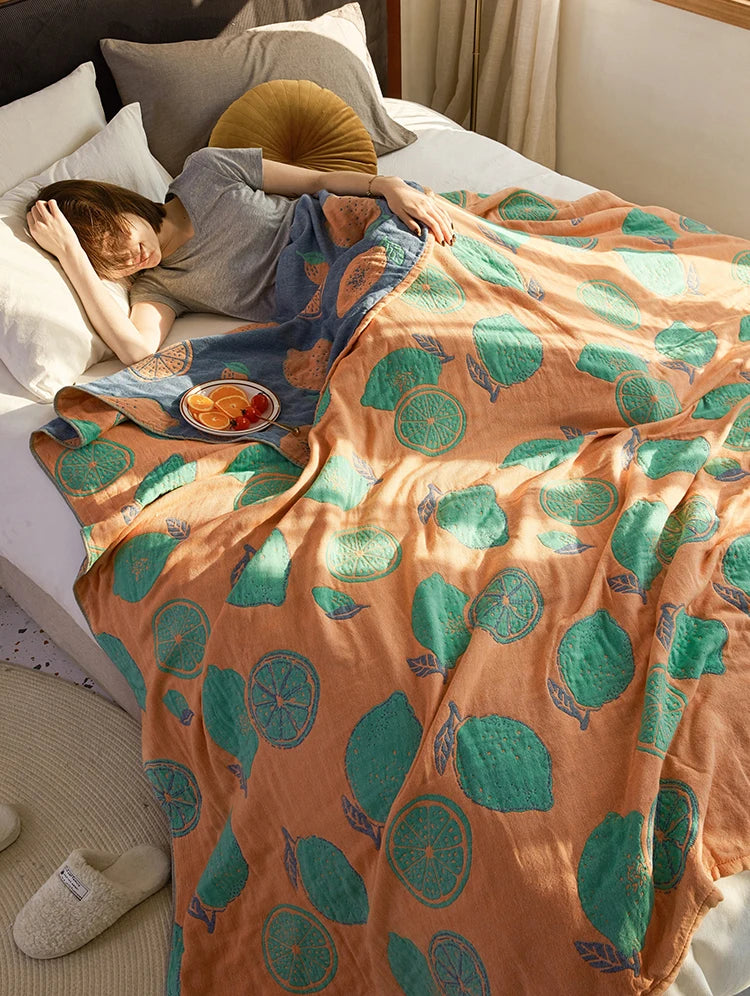 Person lying on a bed under a colorful blanket with fruit pattern. Bohemian cotton gauze quilted bedspread in 4-layer muslin, breathable summer blanket for bed or sofa, lightweight soft throw in assorted colors and patterns. My Boho Home. 