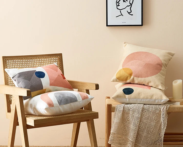 Decorative pillows on a chair and bench against a beige wall with a framed picture. Colorful abstract geometric embroidered cushion cover in cotton blend, decorative pillowcase for living room, kids’ room, or boho home decor.