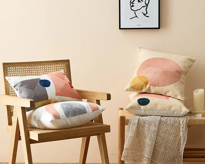 Decorative pillows on a chair and bench against a beige wall with a framed picture. Colorful abstract geometric embroidered cushion cover in cotton blend, decorative pillowcase for living room, kids’ room, or boho home decor.