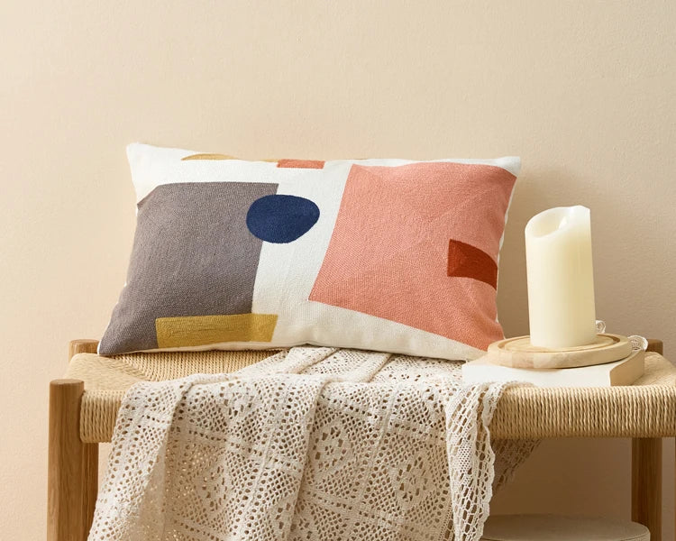 Decorative pillow with geometric pattern on a wooden bench with a candle and blanket. Colorful abstract geometric embroidered cushion cover in cotton blend, decorative pillowcase for living room, kids’ room, or boho home decor.