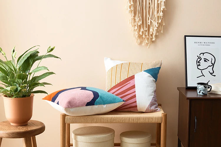 Colorful pillows on a wooden bench with a plant and framed picture in the background Colorful abstract geometric embroidered cushion cover in cotton blend, decorative pillowcase for living room, kids’ room, or boho home decor.