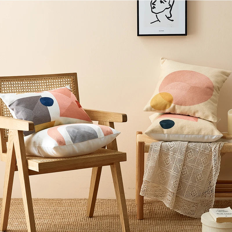 Decorative pillows on a wooden chair and bench against a beige wall. Colorful abstract geometric embroidered cushion cover in cotton blend, decorative pillowcase for living room, kids’ room, or boho home decor.