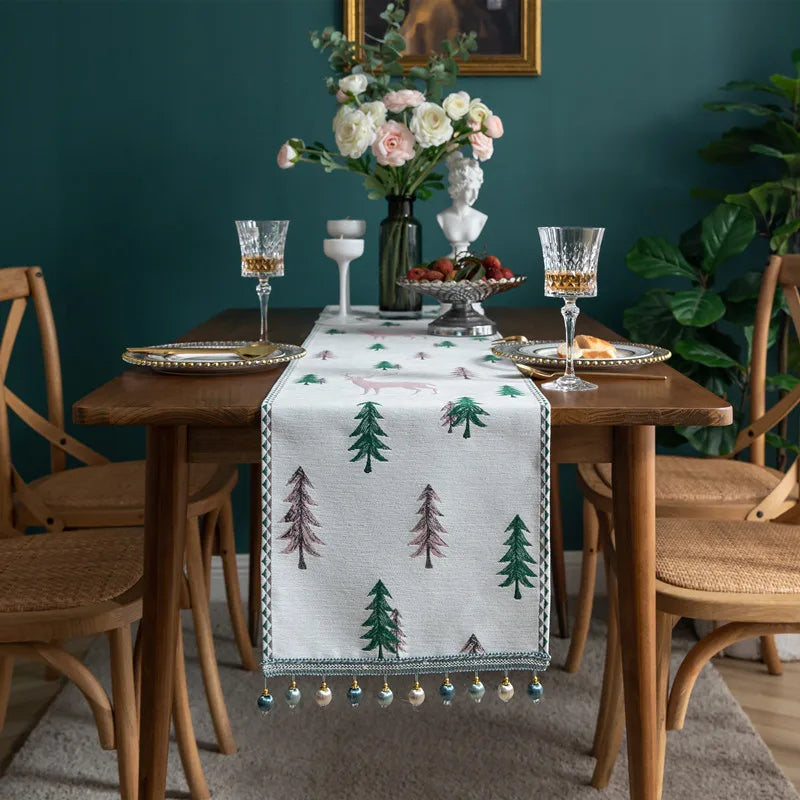 Dining table set with a decorative table runner featuring trees, glasses, and flowers against a teal wall.