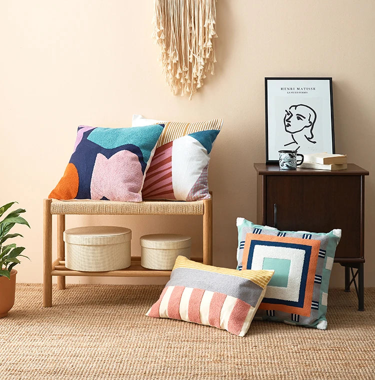 Colorful pillows on a wooden stool against a beige wall with a framed picture. Colorful abstract geometric embroidered cushion cover in cotton blend, decorative pillowcase for living room, kids’ room, or boho home decor.