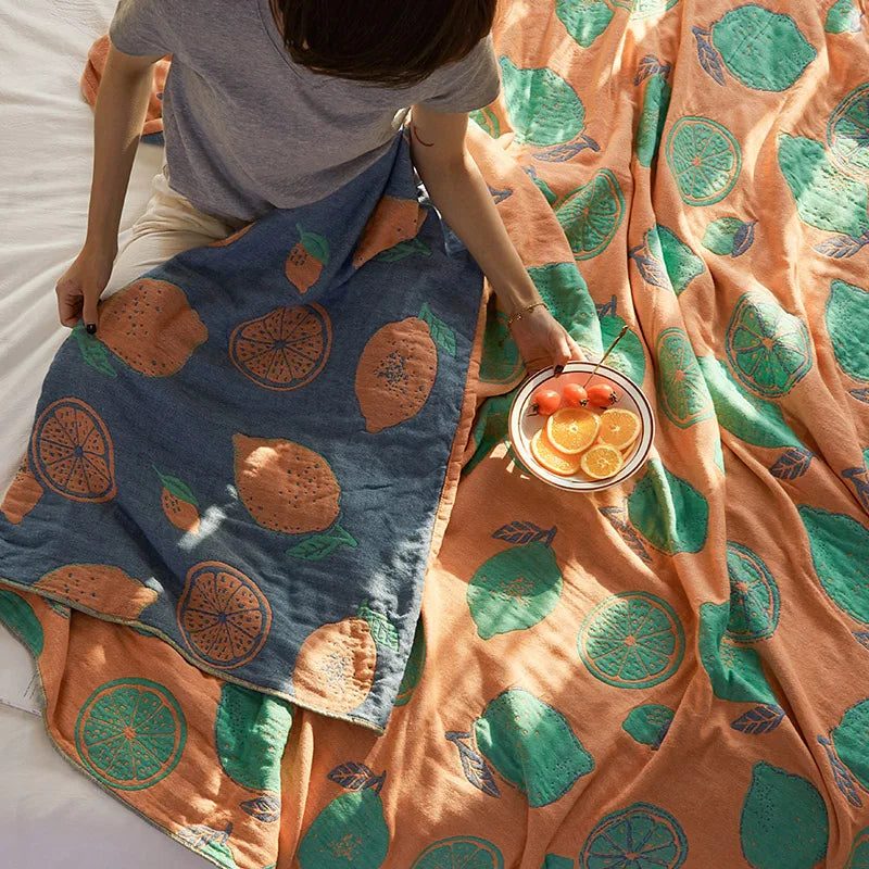Person holding a bowl of fruit next to patterned blankets with orange and green designs. Bohemian cotton gauze quilted bedspread in 4-layer muslin, breathable summer blanket for bed or sofa, lightweight soft throw in assorted colors and patterns. My Boho Home. 