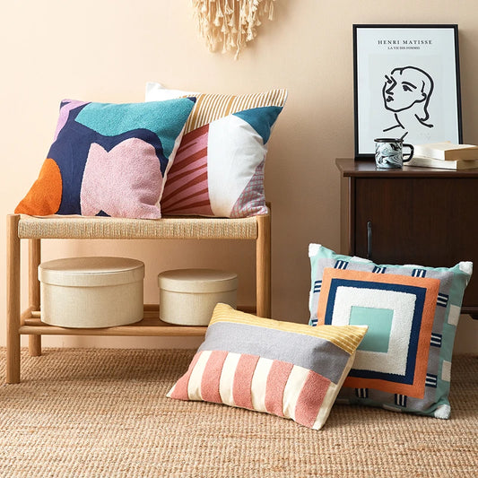 Colorful pillows on a wooden bench and floor in a room with a framed picture on the wall. Colorful abstract geometric embroidered cushion cover in cotton blend, decorative pillowcase for living room, kids’ room, or boho home decor.