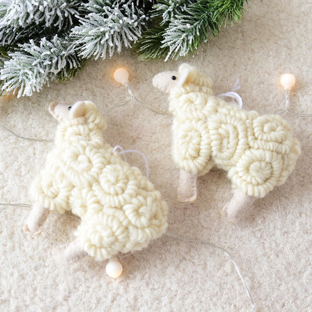 Two white knitted sheep ornaments on a textured surface with Christmas lights and greenery.