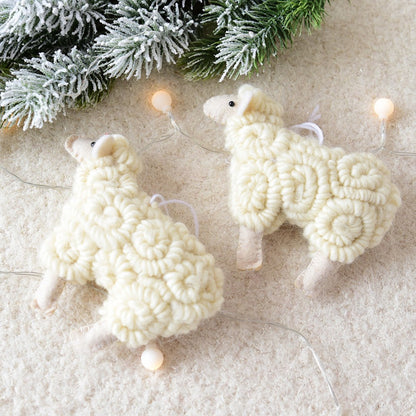 Two white knitted sheep ornaments on a textured surface with Christmas lights and greenery.