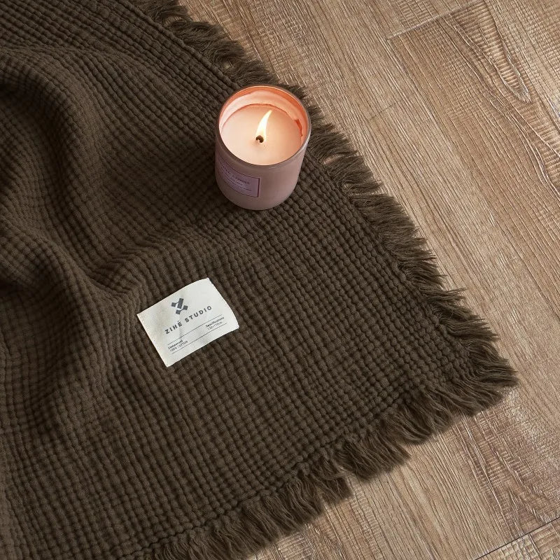 Brown textured blanket with a lit candle and label on a wooden floor Maillard cotton gauze blanket with tassels, four-layer muslin throw for sofa, bed, and boho home decor