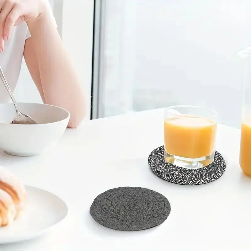 Gray woven coasters on a white surface with a glass of orange juice and a bowl. Set of cotton drink coasters – eco-friendly housewarming gift idea