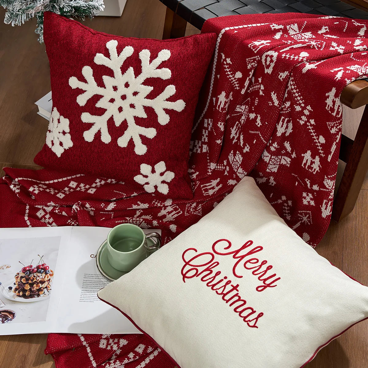 Red and white Christmas-themed pillow and blanket on a chair with a magazine and mug. Embroidered Merry Christmas pillow cover with snowflake design – festive red and white holiday cushion for sofa or bedroom décor.