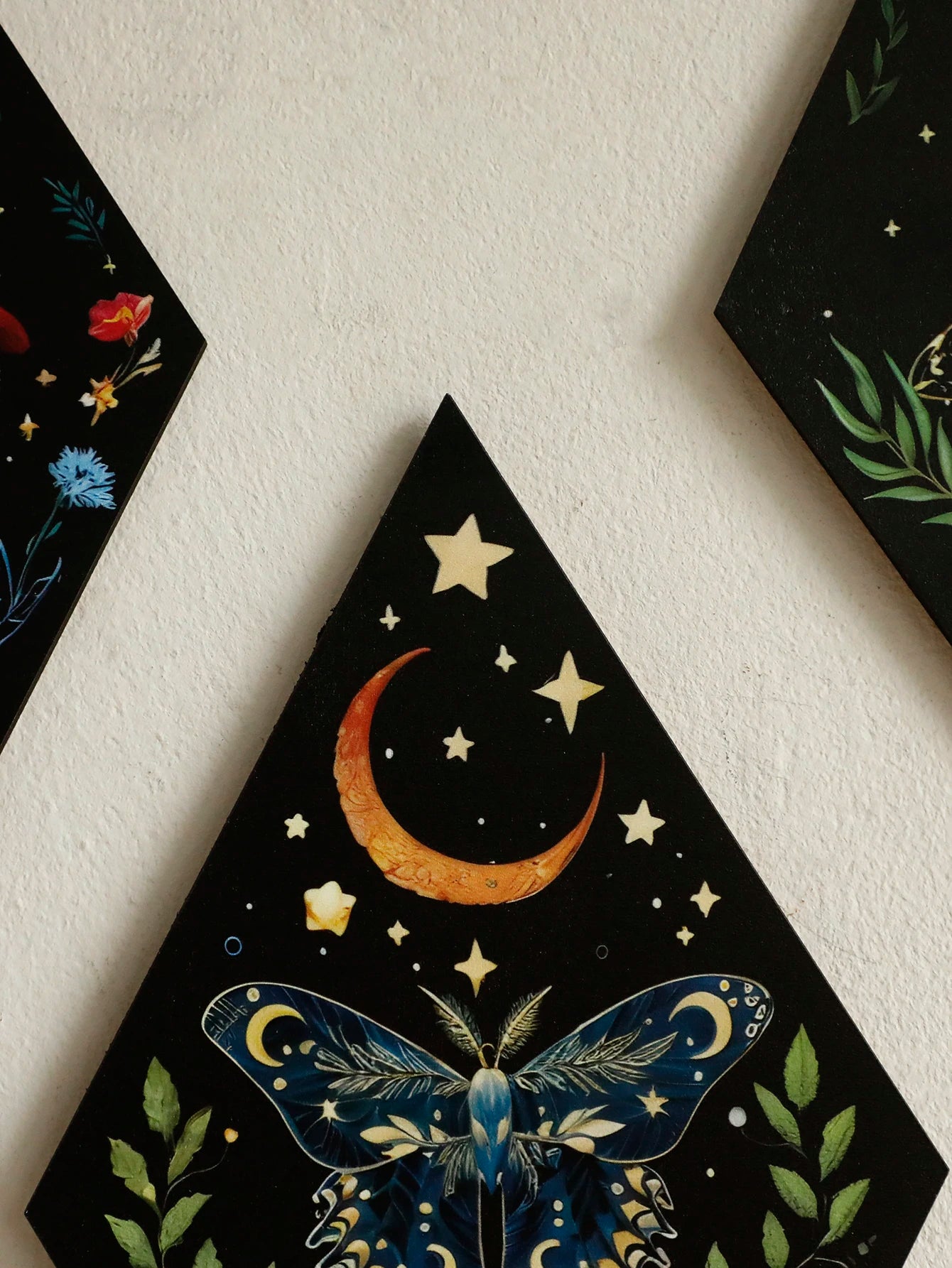 Decorative triangle with celestial design featuring a butterfly, moon, and stars on a black background. Rhomboid-shaped boho wooden wall hanging with engraved celestial patterns, displayed in modern minimalist room