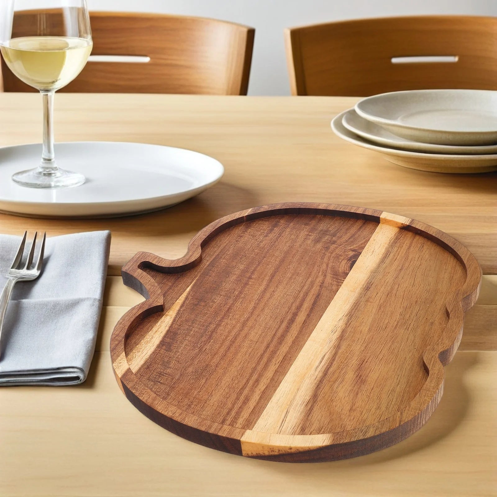 Wooden cutting board on a dining table with plates and a glass of wine. Rustic wooden pumpkin serving platter with natural grain, charcuterie board for Thanksgiving snacks, Halloween desserts, and fall entertaining