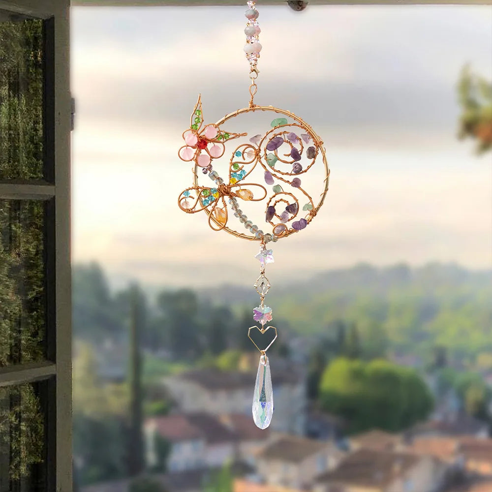 Decorative hanging ornament with flowers and a teardrop-shaped crystal in front of a window with a view of trees and houses. butterfly flower boho home decor, crystal wind chime, sun catcher for windows, moon prism chime, agate stone hanging, garden art decor, patio hanging decoration, boho housewarming gift, spiritual gift, crystal suncatcher, balcony decor, meditation decor, handmade crystal chime