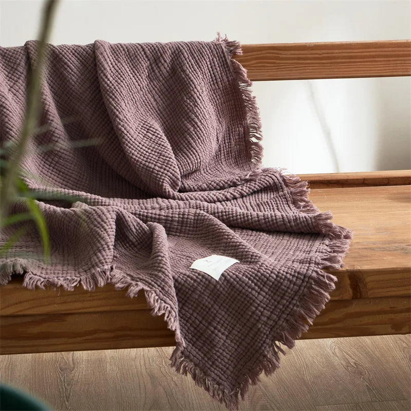 Pink textured blanket draped over a wooden surface with a white label. Maillard cotton gauze blanket with tassels, four-layer muslin throw for sofa, bed, and boho home decor