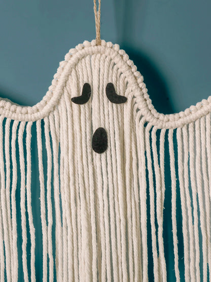 A cream macrame ghost wall hanging with a black felt face, shown hanging against a teal-blue wall from My Boho Home