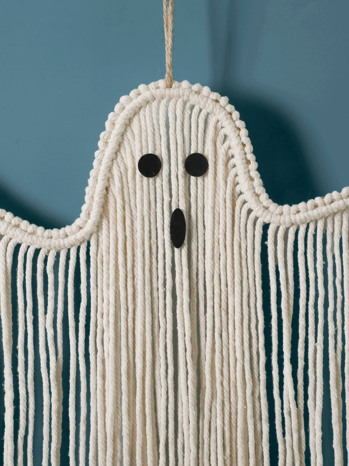 A ghost-shaped macrame wall hanging made of off-white cotton cord, with a simple black face and long fringe, shown against a muted blue background from My Boho Home