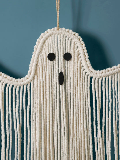 A ghost-shaped macrame wall hanging made of off-white cotton cord, with a simple black face and long fringe, shown against a muted blue background from My Boho Home